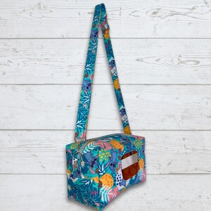 May include: A teal and pink fabric shoulder bag with a zipper closure and a matching strap. The bag features a colorful pattern of cartoon characters, pineapples, and tropical leaves. The bag has a rectangular shape with a clear window.