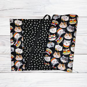 May include: A black book cover with a paw print pattern and a rainbow cat design. The cover is made of fabric and has a black strap.