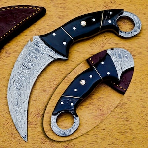 May include: Two karambit knives with curved blades and black handles. The blades have a patterned design, and the handles feature white dots and gold accents. One knife is partially sheathed in a brown leather case.