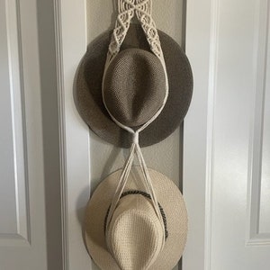 May include: Two straw hats hanging on a macrame hat rack. The hats are brown and beige with a woven texture. The macrame rack is made of white cotton cord and has a simple design.