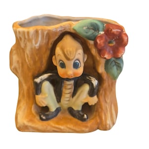 May include: Vintage ceramic planter shaped like a tree stump, featuring a seated cartoon figure. The figure has blue eyes, blonde hair, and is wearing a black jacket and yellow pants. A red flower and green leaves adorn the side.