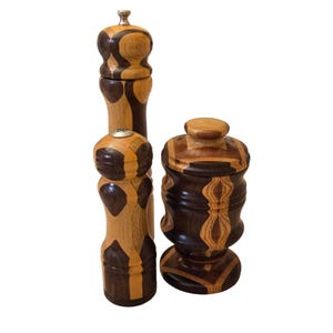 May include: Two tall wooden pepper mills and one shorter wooden lidded container, all featuring a segmented design with alternating light tan and dark brown wood grain patterns. The mills have metallic tops, while the container has a rounded lid.