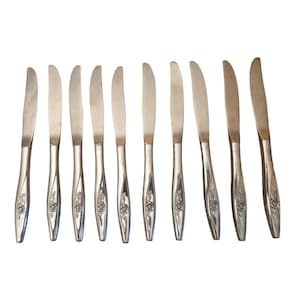 May include: A set of ten silver-toned stainless steel dinner knives arranged in a row. Each knife features a smooth, curved handle with a detailed embossed floral rose motif near the base and a polished, slightly serrated blade edge.
