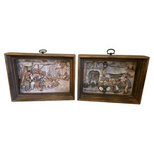 May include: Two rectangular wall plaques featuring three-dimensional relief scenes of outdoor market stalls with figures and produce. Each piece is encased in a dark wood frame with a small metal hanging loop attached to the top center edge.