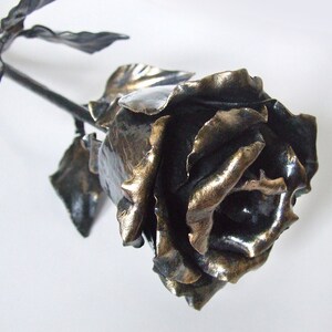 May include: A handcrafted metal rose sculpture with a dark bronze finish. The rose features detailed petals and leaves, with a stem extending from the flower. The sculpture is a decorative item, suitable for display.