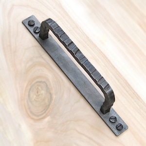 Puede incluir: Una manija de puerta de metal negro con un acabado texturizado y dos tornillos. La manija está diseñada para un cajón o un armario.