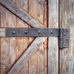 Puede incluir: Primer plano de una puerta de madera rústica con una bisagra de metal negro. La bisagra tiene remaches decorativos y un diseño retorcido.