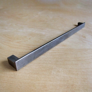 May include: A long, rectangular, metal drawer pull with a brushed finish. The pull is made of a dark gray metal and has a slightly textured surface.