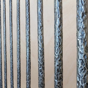 May include: Close-up of several dark gray, textured metal rods. The rods have a hammered appearance, with irregular surfaces. The rods are arranged vertically, suggesting a railing or decorative element. The background is a light beige color.