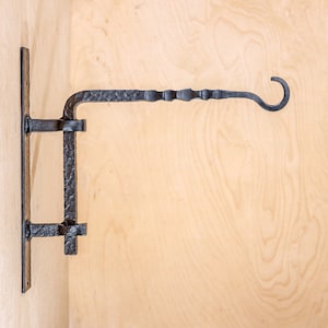 May include: Black metal wall-mounted hook with a decorative scroll design. The hook is attached to a wooden surface.