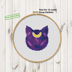 Può includere: Schema a punto croce di una testa di gatto con una luna crescente, nei toni del viola e dell'oro. Il disegno è in un telaio da 15 cm. Il testo "PDF PATTERN" è in un rettangolo verde.