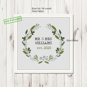 Peut inclure: Un motif de point de croix encadré sur fond blanc. Le motif présente une couronne verte entourant le texte "MR & MRS WILLIAMS est. 2020". Le cadre est blanc, et les dimensions sont de 25,4 cm sur 25,4 cm.