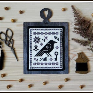 May include: Black and white cross-stitch embroidery of a crow with a key, flowers, and geometric patterns on a white background. The embroidery is framed in a dark wood frame with a heart-shaped top.