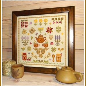 Puede incluir: Un bordado de punto de cruz enmarcado que presenta una tetera, tazas de té y otros motivos florales y geométricos en tonos marrones, amarillos, rojos y verdes. El bordado está hecho sobre un fondo de color crema.