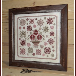 Può includere: Un ricamo a punto croce incorniciato che presenta un bouquet di fiori rossi in un vaso marrone con l'anno 2017 ricamato sotto. Il disegno è circondato da una varietà di forme geometriche rosse, verdi e marroni.