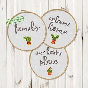 May include: Three round embroidery hoops with cross-stitch designs. Each hoop features a different phrase: "family," "welcome home," and "our happy place." Small cactus designs in orange pots accompany each phrase. The background is a light wood texture.