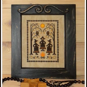 Könnte beinhalten: Ein gerahmtes Kreuzstich-Design mit drei Hexen, die um ein Lagerfeuer versammelt sind, mit einem Vollmond im Hintergrund. Die Hexen tragen schwarze Kleider und spitze Hüte. Das Design ist im rustikalen Stil mit einem schwarzen Rahmen.