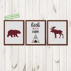 Puede incluir: Conjunto de tres patrones de punto de cruz enmarcados. Uno presenta un oso, otro un alce, y el central dice "little man cave" con un diseño de tipi. Todos los diseños son en cuadros de búfalo rojo y negro sobre fondo blanco.