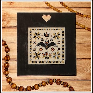 May include: A black framed cross-stitch with a white background featuring a black bat, orange pumpkins, and yellow stars. The frame has a heart-shaped cutout at the top.
