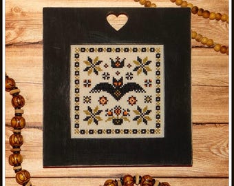 May include: A black framed cross-stitch with a white background featuring a black bat, orange pumpkins, and yellow stars. The frame has a heart-shaped cutout at the top.