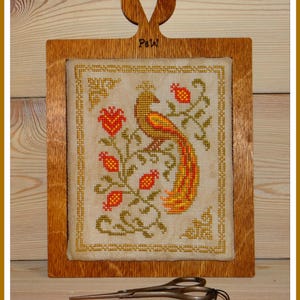 May include: A framed cross-stitch design featuring a stylised peacock with a long tail, surrounded by red and green floral motifs. The frame is made of wood and has a heart-shaped cutout at the top. The text "Paw" is engraved on the frame.