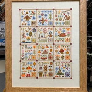 Könnte beinhalten: Ein gerahmter Kreuzstich-Sampler mit 12 verschiedenen Designs, die die Monate des Jahres darstellen. Jedes Design ist nummeriert und zeigt eine Vielzahl von Farben und Mustern.