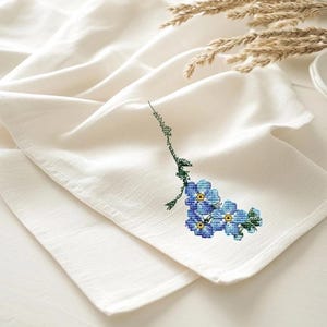 Könnte beinhalten: Ein weißes Küchentuch mit einem gestickten Vergissmeinnicht-Blumendesign in Blau und Grün.