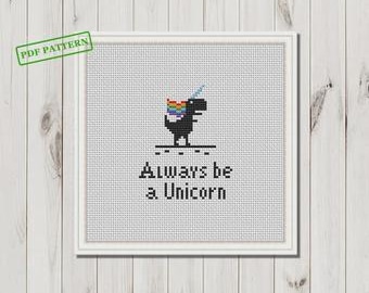 Puede incluir: Un bordado de punto de cruz enmarcado con un dinosaurio negro con melena y cuerno de arcoíris, y el texto "Always be a Unicorn". El marco es blanco y el fondo es un patrón de punto de cruz gris claro.