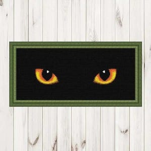May include: Cross-stitch art piece featuring a pair of glowing yellow and orange cat eyes against a black background. The artwork is framed in a green rectangular frame, creating a striking contrast. The piece is displayed on a white wooden surface.