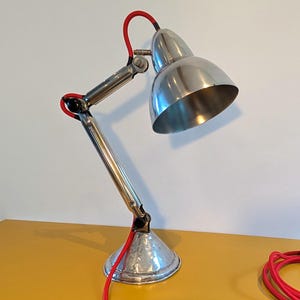 May include: A silver desk lamp with a red cord and a silver shade. The lamp has a flexible arm and a round base.