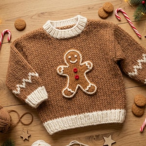 May include: A brown knit sweater featuring a central gingerbread man applique with two red buttons, white ribbed trim at the collar, cuffs, and hem, and white zigzag detailing on the sleeves, crafted from thick yarn for a textured appearance.