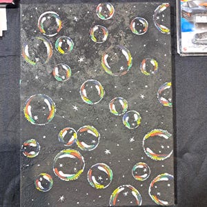May include: An abstract painting on canvas featuring numerous colorful bubbles against a dark gray background. The bubbles have rainbow-colored edges and white highlights, with small white stars scattered throughout the background. The painting is signed "Artzy Henna".