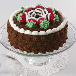 May include: A crocheted chocolate cake with white frosting and red and green strawberries on top. The cake is on a glass cake stand with a white doily underneath.