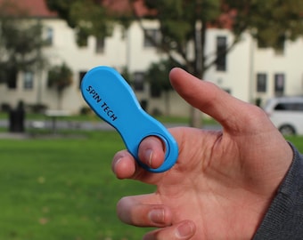 May include: A bright blue, oblong-shaped plastic fidget tool with a circular finger hole at one end. The text "SPIN TECH" is printed in black capital letters on the surface of the blue plastic body, which is held between fingers.