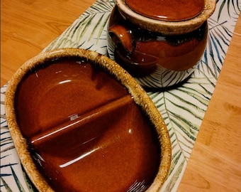 May include: A brown glazed ceramic divided dish and a brown glazed ceramic jar with a lid. The dish and jar are on a table with a green and white patterned tablecloth.