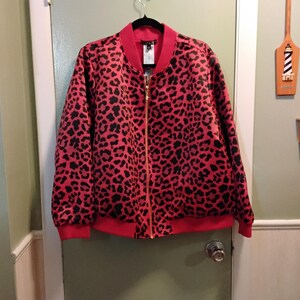 May include: A red bomber jacket with a black leopard print pattern. The jacket has a red collar, cuffs, and hem, and a gold zipper. The jacket is hanging on a black hanger against a green door.