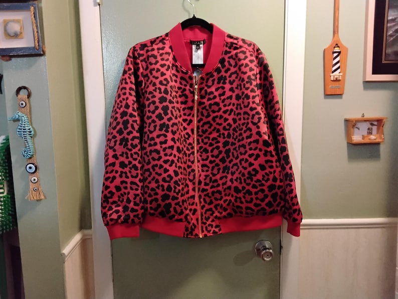 May include: A red bomber jacket with a black leopard print pattern. The jacket has a red collar, cuffs, and hem, and a gold zipper. The jacket is hanging on a black hanger against a green door.