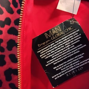 May include: Red and black leopard print fabric with a gold zipper. A black tag with the text "IMAN GLOBAL CHIC" is attached. The tag includes text about fashion and accessories.