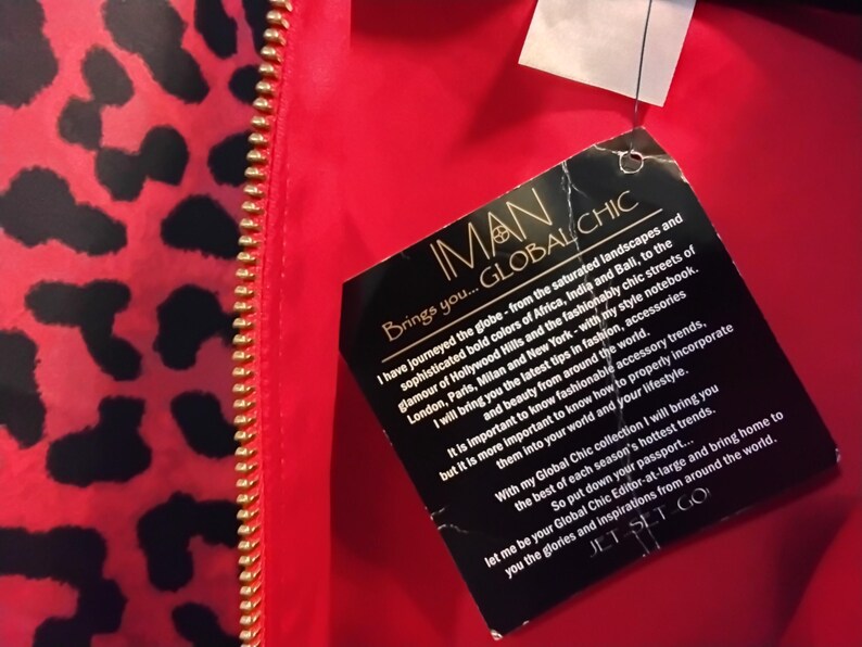 May include: Red and black leopard print fabric with a gold zipper. A black tag with the text "IMAN GLOBAL CHIC" is attached. The tag includes text about fashion and accessories.