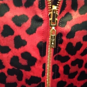 May include: Close-up of a red fabric with black leopard spots. A gold zipper runs vertically down the center, with the brand name "IMAN" on the zipper pull. The fabric has a soft, textured appearance.