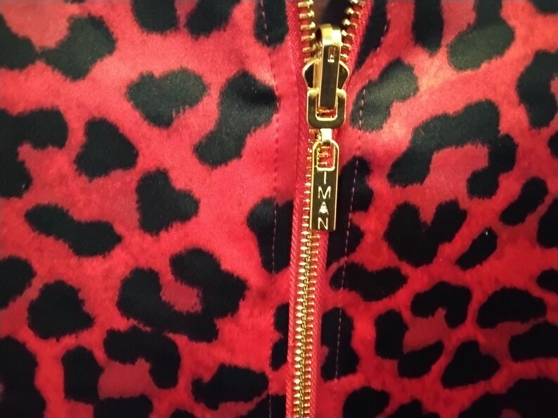 May include: Close-up of a red fabric with black leopard spots. A gold zipper runs vertically down the center, with the brand name "IMAN" on the zipper pull. The fabric has a soft, textured appearance.