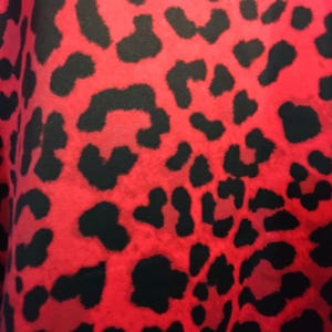 May include: Close-up of a fabric with a vibrant red background and black leopard spots. The pattern is bold and eye-catching, creating a striking visual texture. This textile is suitable for various fashion and design applications.