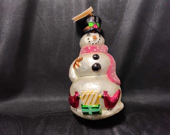 May include: A white glass snowman ornament with a black top hat, pink polka dot scarf, and two red cardinals holding a green and white striped gift box.