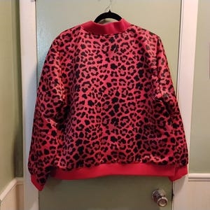 May include: A red and black leopard print sweatshirt with a crew neck and ribbed cuffs and hem. The sweatshirt is hanging on a black hanger against a white door.
