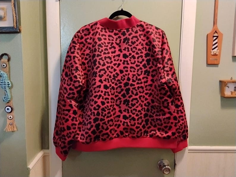 May include: A red and black leopard print sweatshirt with a crew neck and ribbed cuffs and hem. The sweatshirt is hanging on a black hanger against a white door.