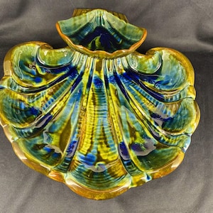 May include: A ceramic seashell-shaped dish with a glossy finish. The dish features a vibrant color palette of yellow, blue, and green, with a brown rim. The design includes a small, raised compartment at the top. This decorative piece is suitable for serving or display.