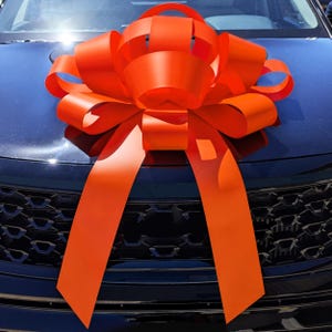 May include: A large, bright orange car bow is attached to the hood of a dark blue vehicle. The bow is made of wide, folded ribbons, creating a full, decorative shape. Long, flowing ribbons extend from the bow, adding to its festive appearance.
