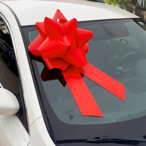 May include: A white car with a large red bow on the windshield. The bow is tied with a long red ribbon.