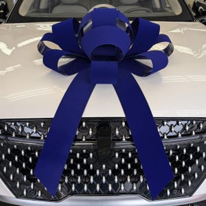 May include: A large, navy blue bow adorns the hood of a white car. The bow is made of wide, velvety ribbon and is centered above the car's grill. The bow's long streamers cascade down the front of the vehicle.