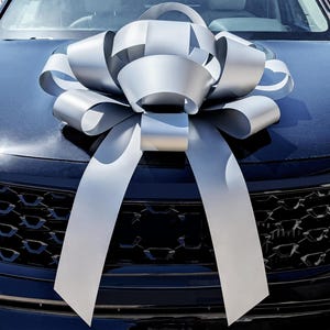 May include: A large, silver bow adorns the hood of a black car. The bow is made of wide, metallic ribbon, with long, flowing tails. The bow is centered on the hood, above the car's grill.